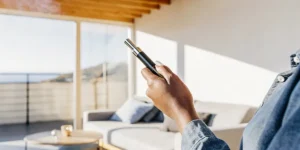 A person's hand holding a stylish vape pen indoors, with a bright window and coastal view in background.