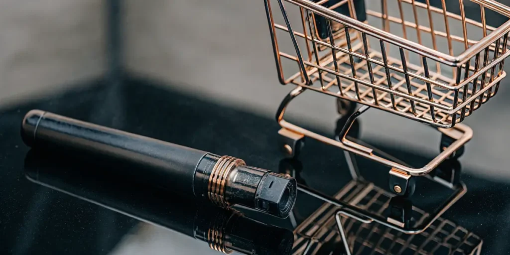 Artistic macro shot: black vape pen lying next to a miniature golden shopping cart on a reflective surface.