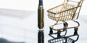 Artistic macro shot: gold vaporizer standing next to a miniature golden shopping cart on a reflective surface.