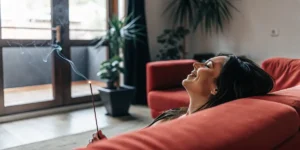 Woman lounging on a red sofa, eyes closed, smiling while holding a smoking incense stick in a modern living room.