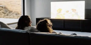 Couple relaxing on a couch, watching an animated show on a wall-mounted TV in a cozy, sunlit living room.